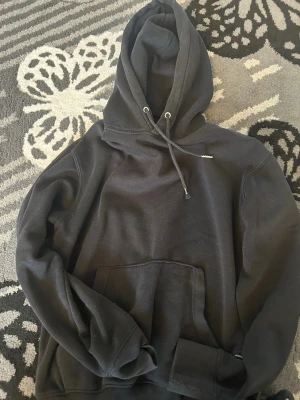 Svart hoodie med ficka och huva - Säljer en svart hoodie med stor känguruficka framtill och justerbar huva med dragsko. Hoodien har en avslappnad passform och ribbade muddar vid ärmar och nederkant. Perfekt för en chill och stilren look.