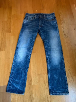 Blå raka jeans med slitningar - Snygga blå jeans med raka ben och tydliga slitningar framtill och baktill. Jeansen har klassisk femficksdesign och är tillverkade i denim. Perfekta för en avslappnad och trendig stil.