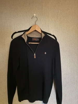 Svart half zip-tröja från Morris - Klassisk svart tröja från Morris med half zip och liten logga på bröstet. Tröjan har en ståkrage med beige insida och är tillverkad i mjukt material. Perfekt för en stilren och avslappnad look.