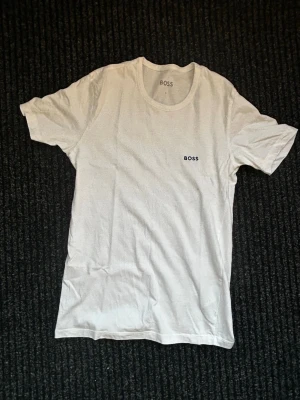 Vit t-shirt från BOSS - Snygg vit t-shirt från BOSS med diskret logga på bröstet. Klassisk rund halsringning och korta ärmar. Perfekt basplagg som funkar till allt och har en clean look. Tillverkad i mjuk bomull för skön känsla hela dagen.