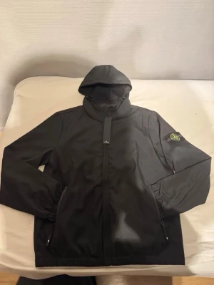 Svart vindjacka från Stone Island - Snygg svart vindjacka från Stone Island med ikonisk patch på vänster ärm. Jackan har huva, hel dragkedja framtill och två fickor med dragkedja. Tillverkad i ett lätt och slitstarkt material som passar perfekt för blåsiga dagar.