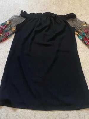 Svart klänning med broderade meshärmar - Svart axelbandslös klänning med raka linjer och transparenta meshärmar. Ärmarna har färgglada broderade blommor i rött, turkost och gult. Klänningen har en elastisk kant upptill för en snygg off-shoulder-look. Perfekt för dig som gillar unika detaljer.
