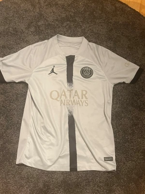 PSG matchtröja vit Jordan x Nike - Snygg vit PSG matchtröja med svarta detaljer och logga från Jordan och Paris Saint-Germain på bröstet. Tröjan har svart rand i mitten, Qatar Airways-tryck och svarta muddar. Perfekt för fotbollsträning eller att bära som supporter. Tillverkad i lätt och ventilerande material.