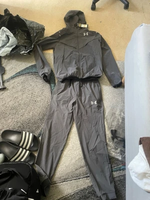 Grå träningsset från Under Armour - Säljer ett grått träningsset från Under Armour med jacka och byxor. Setet är gjort i ett lätt och funktionellt material, perfekt för träning eller uppvärmning. Jackan har huva och dragkedja, och båda plaggen har logga på bröst och ben.