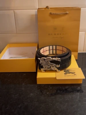 Burberry bälte - Snyggt svart bälte från Burberry med klassiskt rutigt mönster på insidan och en stor silverfärgad spänne med Burberrys ikoniska riddare. Perfekt accessoar för att lyfta din outfit.