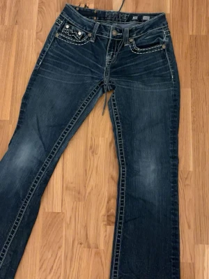 Mörkblå bootcut jeans från Miss Me - Snygga mörkblå jeans från Miss Me med bootcut passform och markerade vita sömmar. Jeansen har klassisk femficksdesign, låg midja och slitningar på låren för en cool look. Perfekta för dig som gillar detaljer och streetstyle.