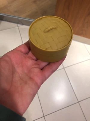 Gul rund liten förvaringsburk med mystery dumpling innuti  - Säljer en liten rund förvaringsburk i gul plast med lock som har ett flätat mönster och en liten detalj på toppen. Perfekt för småsaker eller smycken. Enkel och stilren design som passar i alla rum.
