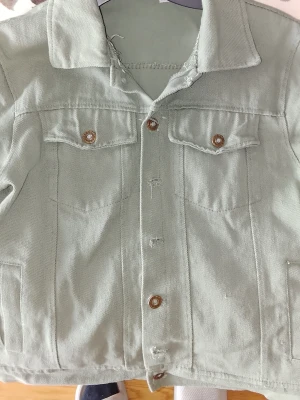 Ljusgrön jeansjacka med bruna knappar - Snygg ljusgrön jeansjacka med klassisk krage och bruna knappar framtill. Jackan har två bröstfickor med lock och knapp samt två sidofickor. Rak passform och stilren design, perfekt för en trendig look. Waist-size, knappast använt den pga för liten, inga fläckar eller repor.
