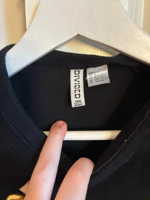Svart ribbad croppad topp H&M Divided - Svart croppad topp från H&M Divided med ribbad struktur och korta ärmar. Enkel och stilren design som passar perfekt till jeans eller kjol. Mjuk och stretchig för en bekväm passform.