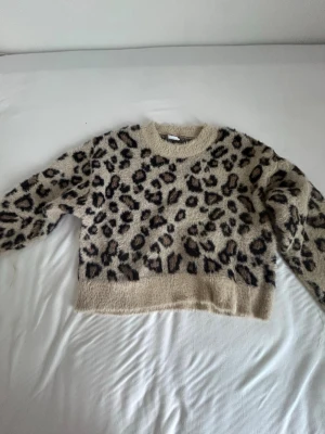 Beige leopardmönstrad stickad tröja - Mysig stickad tröja i beige med leopardmönster i svart och brunt. Tröjan har rund halsringning, långa ärmar och en fluffig, mjuk känsla. Perfekt för dig som vill sticka ut med ett djurmönster i garderoben.Den har några snören som sticker ut.