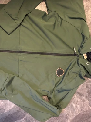 Grön vindjacka från Moncler - Snygg grön vindjacka från Moncler med svart dragkedja och Moncler-logga på bröstet. Jackan har en minimalistisk design och är tillverkad i ett lätt material som passar perfekt när det blåser. Praktisk huva och diskreta detaljer gör den enkel att matcha.