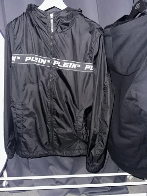 ÄKTA Svart vindjacka från Philipp Plein - Svart vindjacka från Philipp Plein med vit logga längs bröstet. Jackan har huva, dragkedja framtill och sidofickor. Tillverkad i lätt och glansigt material med meshfoder på insidan. Perfekt för en sportig och trendig look. ÄKTA Kvitto finns vid förfrågan