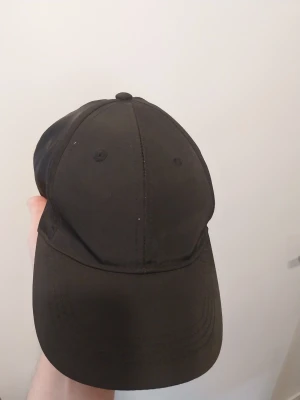 Svart klassisk keps med justerbart spänne - En stilren svart keps i klassisk modell med böjd skärm och justerbart metallspänne baktill. Kepsen är enkel och passar till de flesta outfits. Tillverkad i ett mjukt tygmaterial för skön passform.