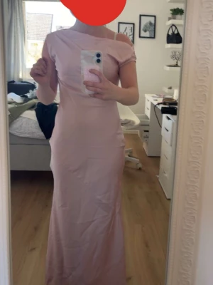 Ljusrosa långklänning med asymmetrisk axel - Elegant ljusrosa långklänning med asymmetrisk axel och kort ärm. Klänningen har en smickrande, figurnära passform och är tillverkad i ett mjukt, slätt material. Perfekt för speciella tillfällen när du vill sticka ut med en stilren look. Perfekt till balen eller en festlighet. 