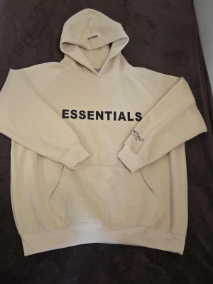 Essentials hoodie beige - Använd en gång. Snygg beige hoodie från Essentials med svart tryck på bröstet och ärmen. Klassisk känguruficka framtill och stor huva. Perfekt för en avslappnad streetwear-look. Mjuk bomullsblandning som känns skön mot huden.