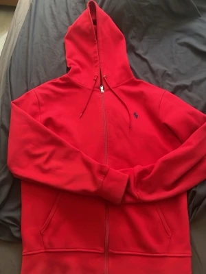 Röd hoodie från Polo Ralph Lauren - Riktigt snygg röd hoodie från Polo Ralph Lauren med dragkedja, huva och dragsnören. Klassisk blå logga broderad på bröstet. Mjuk bomullsblandning och två fickor framtill. Perfekt för en chill och stilren look.