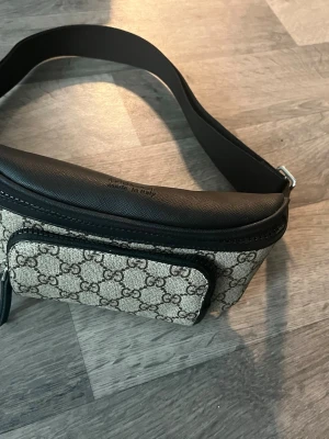 väska - snygg midjeväska i beige canvas med klassiskt GG-mönster och svarta detaljer i läder. Väskan har ett justerbart axelband och en praktisk ytterficka med dragkedja. Perfekt för dig som vill ha en trendig och lyxig accessoar.