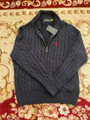 Mörkgrå kabelstickad tröja Polo - Snygg mörkgrå kabelstickad tröja från Polo Ralph Lauren med halv dragkedja och hög krage. Tröjan har det klassiska röda Polo-logot broderat på bröstet och ribbstickade muddar. Perfekt för en stilren och avslappnad look.