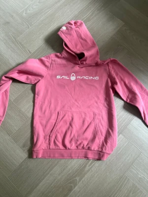 Rosa hoodie från Sail Racing - Säljer en snygg rosa hoodie från Sail Racing med vit logga på bröstet. Hoodien har huva, magficka och ribbade muddar vid ärmslut och nederkant. Perfekt för en avslappnad och sportig stil.