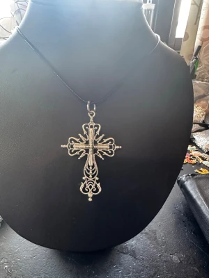 Silvrigt halsband med korshänge - Snyggt halsband med ett stort, detaljerat korshänge i silverfärgad metall. Hänget har en dekorativ och vintageinspirerad design med fina mönster och små kulor. Hängs på en tunn svart snodd som ger en cool kontrast till det silvriga hänget.