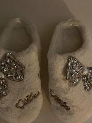 Vita fluffiga tofflor med strassrosett, storlek 36-37 - Supermjuka och fluffiga vita tofflor med en dekorativ rosett med glittrande strass – både bekväma och snygga! Perfekta för mys hemma! Storlek: 36–37. Säljs i begagnat skick! Kika gärna in på mina andra annonser och hör av dig vid frågor eller intresse!