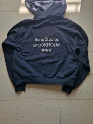 Svart hoodie Acne Studios Stockholm - Svart hoodie från Acne Studios med tryck på ryggen där det står 'Acne Studios STOCKHOLM 1996' i vitt. Klassisk modell med huva och ribbade muddar. Perfekt för dig som gillar stilrena och trendiga plagg med streetkänsla.