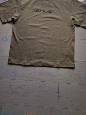 Beige Acne Studios t-shirt med pärlor - Snygg beige t-shirt från Acne Studios med coolt pärlbroderat logotyptryck på bröstet. Klassisk rund halsringning och korta ärmar. Perfekt för dig som gillar stilrena plagg med en twist.