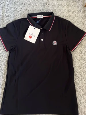 Svart pikétröja från Moncler - Svart pikétröja från Moncler med klassisk krage och korta ärmar. Tröjan har vita och röda detaljer på krage och ärmslut samt Moncler-logga på bröstet. Perfekt för en stilren och sportig look.