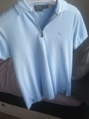 Ljusblå polotopp från Ralph Lauren - Knappt använd Snygg ljusblå polotopp från Ralph Lauren med kort ärm och halv dragkedja framtill. Klassisk krage och broderad logga på bröstet. Modellen är slim fit och tillverkad i stretchig bomullsmix för en bekväm passform.