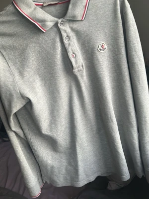 Grå långärmad polotopp från Moncler - Snygg grå långärmad polotopp från Moncler med klassisk krage och knappar framtill. Diskret Moncler-logga på bröstet och röda/vita detaljer på krage och ärmslut. Perfekt för en stilren och sportig look.