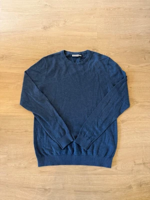 Mörkblå rundhalsad tröja från kappahl - En stilren mörkblå tröja från kappahl med rund halsringning och långa ärmar. Tröjan har ribbade muddar vid ärmslut och nederkant, perfekt för att hålla värmen. Materialet är mjukt och tunt, vilket gör den skön att bära.