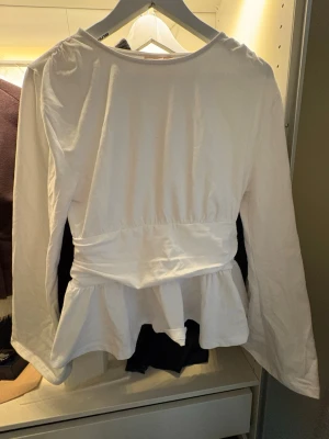 Vit peplumblus från Gina Tricot - Snygg vit blus från Gina Tricot med peplumdetalj i midjan och långa ärmar. Blusen har rund halsringning och är tillverkad i ett mjukt, lätt material som ger en skön känsla. Perfekt för att lyfta din outfit med en stilren touch. Strl 170 men passar S