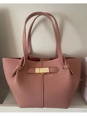 Lindex Rosa handväska med gulddetalj - Söt rosa handväska i skinnimitation med både handtag och justerbar axelrem. Väskan har en dekorativ rosett framtill med guldfärgad detalj. Perfekt storlek för det viktigaste och ger en trendig touch till din outfit.