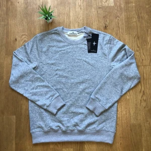 Grå stickad tröja från Stone Island - Snygg grå stickad tröja från Stone Island med rund halsringning och ribbade muddar vid ärmslut och nederkant. Tröjan har en enkel och stilren design som passar perfekt till många olika outfits. Materialet är mjukt och bekvämt.