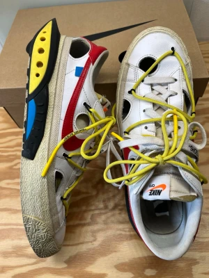 Nike x Off-White Blazer Low - Unika Nike x Off-White Blazer Low sneakers med vit läderöverdel, röda och blå detaljer, samt gula skosnören. Skorna har cut-out hål, Off-White-typisk text och en chunky sula med svarta, gula och blå inslag. Swoosh-logga i rött och Off-White branding på sidan.