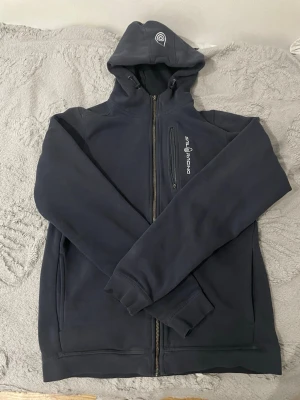 Mörkblå hoodie från Sail Racing - Mörkblå hoodie med dragkedja från Sail Racing. Har en bröstficka med dragkedja och logga tryckt på bröstet samt en broderad logga på huvan. Mjuk insida och ribbade muddar vid ärmslut och nederkant. Perfekt för en sportig och avslappnad stil. Fast en bit av dragkedjan har gått av men funkar jätte bra ändå. 