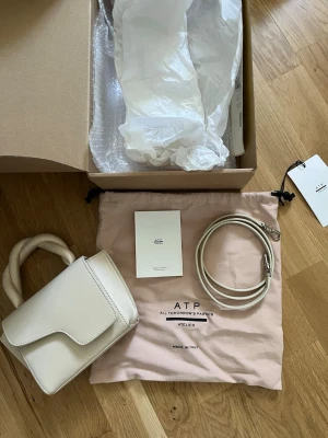 Vit handväska från ATP Atelier - Stilren handväska i ljusbeige läder från ATP Atelier. Väskan har ett unikt lock, tvinnat handtag och medföljande axelrem. Kommer med dustbag och originalförpackning. Perfekt accessoar för en trendig look. Helt ny, använd max 4 gånger. Köpt för ca 1 år sedan. 