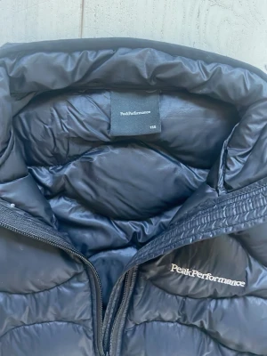 Svart dunväst från Peak Performance - Snygg svart dunväst från Peak Performance med quiltat mönster och hög krage. Västen har dragkedja framtill, två fickor med dragkedja och diskret logga på bröstet. Perfekt för lager-på-lager och håller dig varm under kyliga dagar.