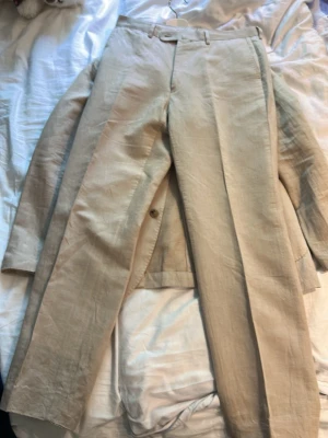 Beige linnékostym med kavaj och byxor - Säljer en stilren beige kostym i linne med klassisk kavaj och matchande byxor. Kavajen har två knappar, utanpåfickor och enkel krage. Perfekt för dig som vill ha en luftig och snygg look till sommaren.