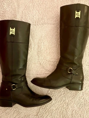 Ralph Lauren klassiska svarta ridstövlar i äkta skinn - 🐎 Lauren Ralph Lauren "Melody" svarta ridstövlar i äkta läder, storlek 38,5. Säljer ett par otroligt snygga och klassiska ridstövlar från Lauren Ralph Lauren i mjukt svart läder. Signaturdetaljer med guldfärgade RLL-logotypbeslag vid skaften och gulddetaljerade selremmar. Stilrent snitt med låg/medel klack. En mycket god investering! Designermärke: Lauren Ralph Lauren. Modell: Melody. Storlek: US 8B / c:a EU 38,5 (notera att jag som 39-bärare tyvärr EJ kunde ha dem). Originalpris: 3.000 kr.
