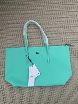 Turkos väska från Lacoste - Snygg turkos shoppingväska från Lacoste med klassisk krokodillogga framtill. Väskan är tillverkad i slitstark PVC och har två långa handtag, perfekt att bära på axeln. Enkel och stilren design med dragkedja upptill och etikett kvar. Ny och helt oanvänd! 