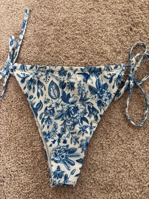 Blåvit blommig bikini med knyt - Bikini med blått blommönster på vit botten. Trekantsmodell upptill och knyt i sidorna på underdelen. Det står att den är i Storlek L men passar mig som har storlek S så skulle säga att det är mer åt storlek M. Ej vaddering. Väldigt bra skick knappt använd.💗