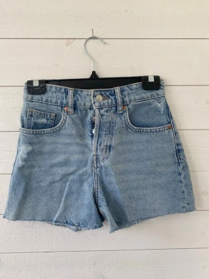 Ljusblå denimshorts - Säljer ett par ljusblå denimshorts med hög midja och rå, fransig kant. Shortsen har klassisk femficksdesign och slitna detaljer framtill för en avslappnad look. Perfekta till sommaren och lätta att matcha med olika stilar.