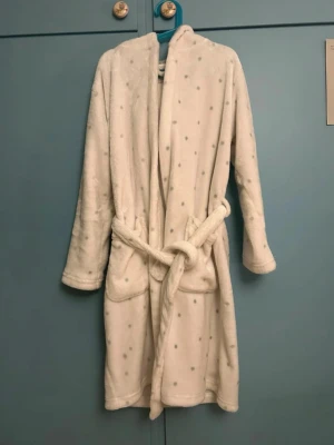 Vit morgonrock med grå prickar - Mysig vit morgonrock med grå prickar och huva. Tillverkad i supermjuk fleece och har knytskärp i midjan. Perfekt för att chilla hemma eller efter duschen. Lång ärm och knälång modell.