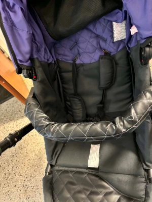 Svart och lila barnvagn med quiltad sits - Barnvagn i svart och lila med quiltad sits och vadderad bygel. Vagnen har justerbart handtag, säkerhetssele och varukorg undertill. Sitsen är mjuk och har detaljer i lila tyg. Passar för promenader och shopping.