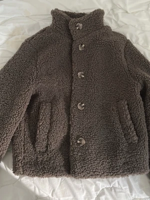 Brun teddyjacka från H&M XS - Mysig brun teddyjacka från H&M i storlek XS. Jackan har hög krage, knäppning med fem stora knappar och två snedställda fickor framtill. Perfekt för kalla dagar och ger en trendig look med sitt fluffiga material.