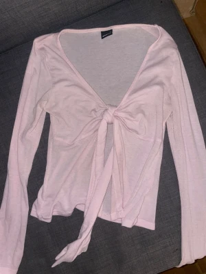 Ljusrosa omlottopp från Gina Tricot - Supersöt ljusrosa omlottopp från Gina Tricot med långa ärmar och knyt framtill. Tunn och mjuk i materialet, perfekt att styla med jeans eller kjol. Djup v-ringning och feminin look.