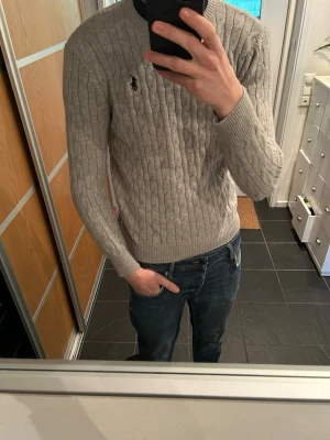 Grå kabelstickad tröja från Polo Ralph Lauren M - Snygg grå kabelstickad tröja från Polo Ralph Lauren med rund halsringning och svart broderad logga på bröstet. Tröjan har långa ärmar och ribbade muddar vid ärmslut och nederkant. Perfekt för lager-på-lager och stilren look.