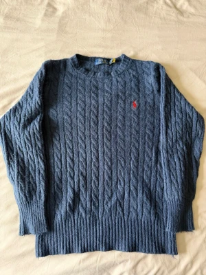Blå kabelstickad tröja från Polo Ralph Lauren - Kabelstickad blå tröja från Polo Ralph Lauren med rund halsringning och röd broderad logga på bröstet. Tröjan har långa ärmar och breda ribbade muddar vid ärmslut och nederkant. Perfekt för lager-på-lager och snygg till jeans.