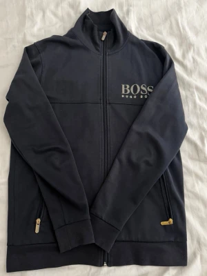 Svart tröja med dragkedja Hugo Boss - Snygg svart hoodie från Hugo Boss med dragkedja framtill och två fickor med guldfärgade dragkedjor. Bra skick och inga problem med den. 
